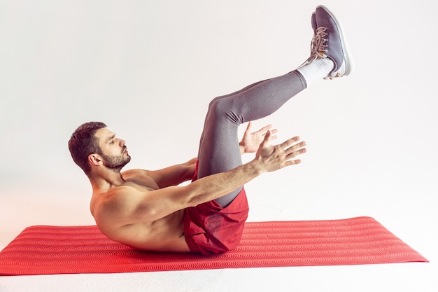 Sportivo europeo che fa scricchiolii per i muscoli addominali sul tappetino fitness. giovane uomo di bell'aspetto concentrato con torso sportivo nudo. isolato su sfondo beige. riprese in studio