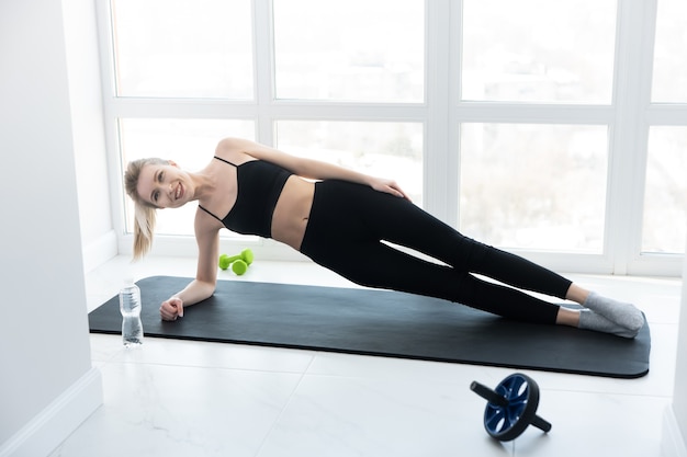 European sportswoman doing exercise on fitness mat. Young beautiful smiling blonde woman wear sportswear. Concept of sport activity at home. Interior of modern spacious apartment. Sunny daytime