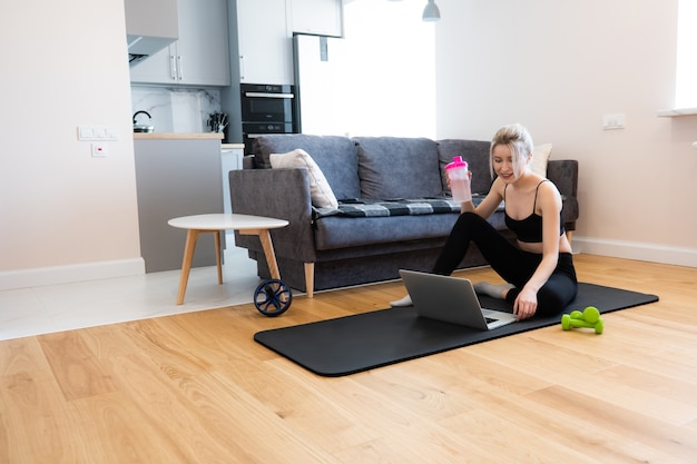 European sportswoman sitting on fitness mat and watching something on laptop computer. young beautiful smiling blonde woman wear sportswear and holding bottle with water. interior of studio apartment