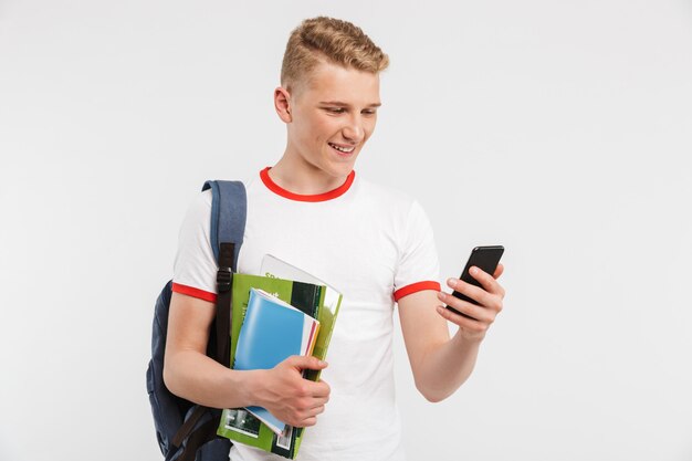 european student boy wearing backpack smiling and using smartphone while holding colorful exercise books isolated on white