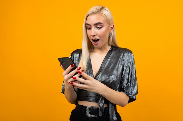 Европейская стильная очаровательная женщина с удивлением читает сообщения на своем смартфоне на желтой стене