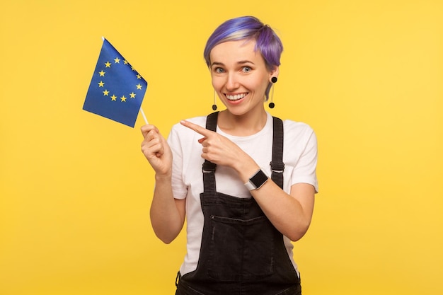 Foto bandiera dell'unione europea ritratto di ragazza hipster positiva con i capelli corti viola in tuta di jeans che punta alla bandiera dell'ue e guardando la fotocamera con un sorriso a trentadue denti isolato su sfondo giallo girato in studio