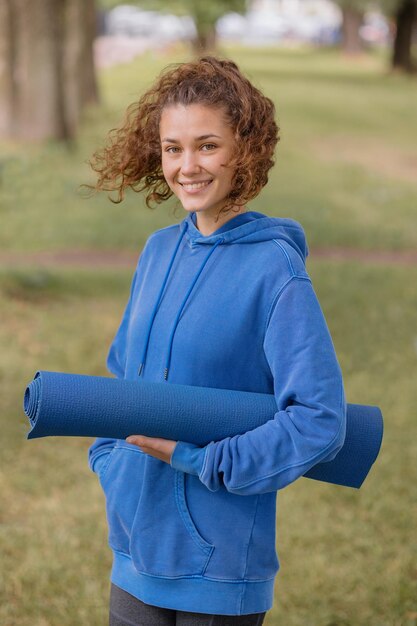 Photo a european woman in a blue hoodie with a blue yoga mat chooses a place to do sports and meditation