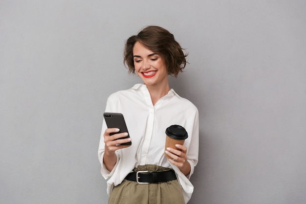 european woman holding takeaway coffee and using cell phone, isolated over gray wall