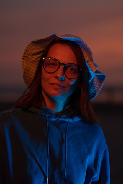 A european woman illuminated by neon light in a hat against the\
background of the sunset sky