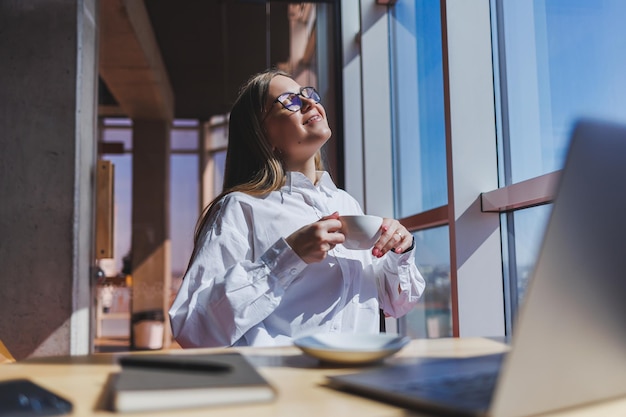 European woman manager sits at a table while working on a laptop and drinks coffee in a cafe A young smiling woman wears glasses and a white shirt Freelance and remote work Modern female lifestyle