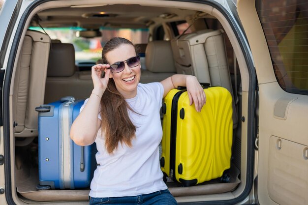 Photo european woman in sunglasses packs her bags for a car trip beautiful happy girl sitting in the trunk of a car and ready for travel yellow suitcase with things for summer vacation