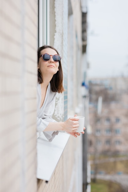 European woman in sunglasses and with a mug of coffee or tea looking out of the window and smiling