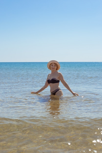 海で水着と腰までの長さの帽子をかぶったヨーロッパの女性が微笑んでカメラをのぞき込む。海辺での休暇。