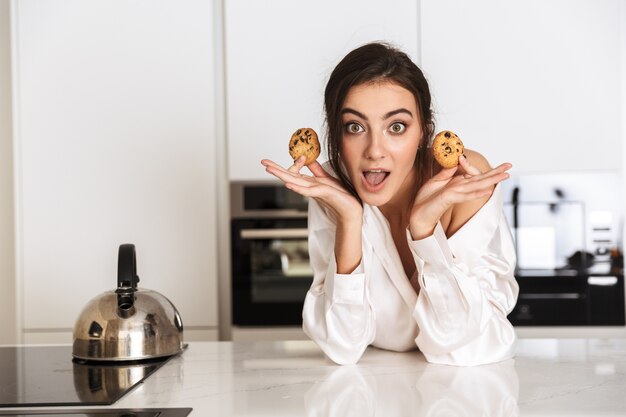 european woman wearing silk clothing holding cookies, while standing in kitchen at home