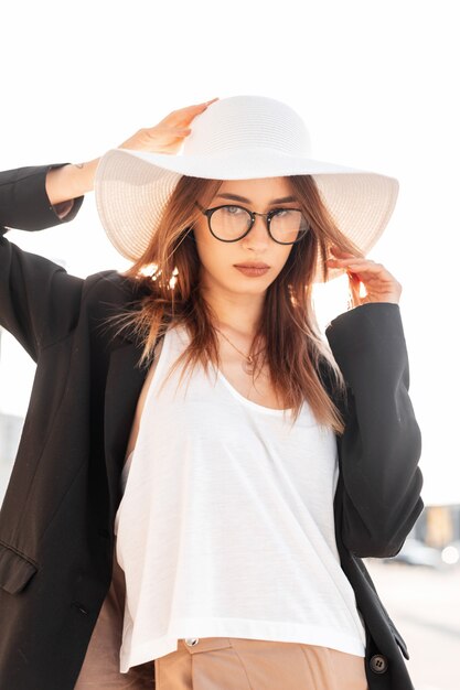 European young beautiful woman in vintage straw hat in stylish glasses in elegant clothes posing on background of bright sunlight on summer day. Sexy urban girl model enjoys sunny day on street.