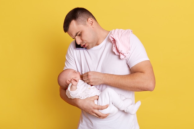 Европейский молодой отец занят телефонным разговором, позирует со своим новорожденным ребенком