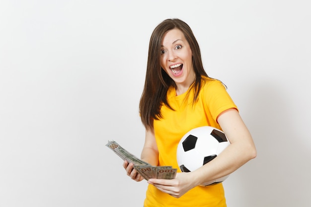 European young fun woman, football fan or player in yellow\
uniform holding bunch of money banknotes, soccer ball isolated on\
white background. sport, play football game, excitement lifestyle\
concept.