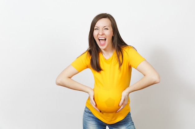 European young happy woman football fan or player with soccer ball under yellow uniform as pregnant with big belly isolated on white background. Sport play football, health, healthy lifestyle concept.