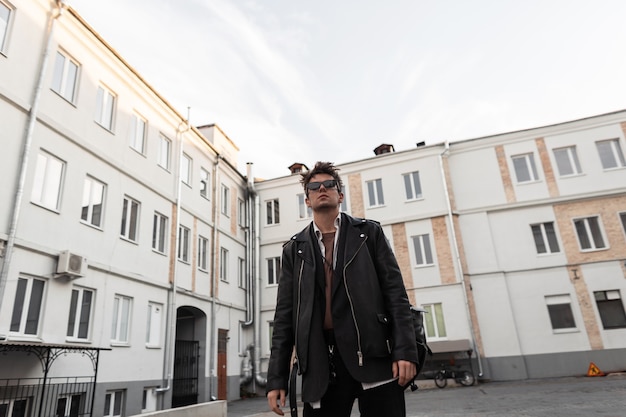 European young man in fashion youth oversized leather clothes with a trendy hairstyle is resting near a modern buildings in the city on a spring day. Modern guy hipster in stylish sunglasses outdoors.