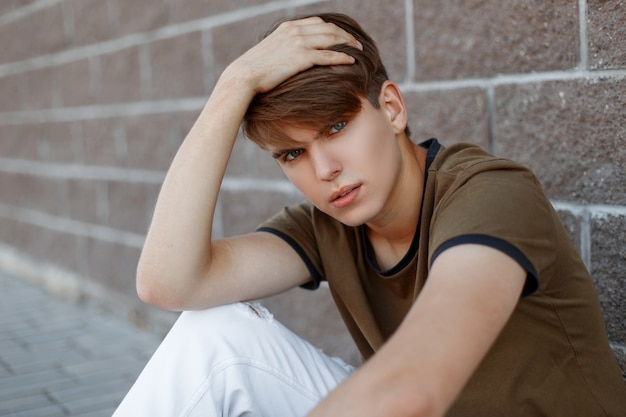 European young man in a fashionable green T-shirt in stylish white pants with a fashionable hairstyle relaxes sitting near a vintage brick wall