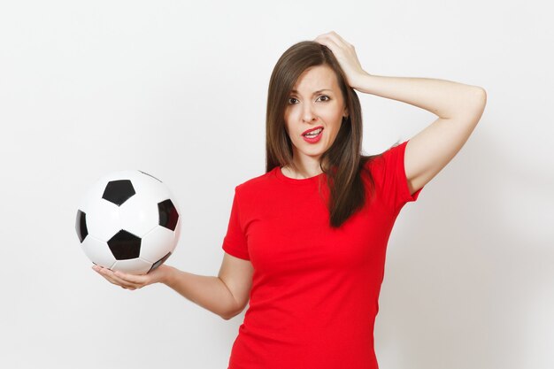 European young sad upset woman, football fan or player in red uniform holds soccer ball, hand on head, worries about losing team isolated on white background. Sport, play football, lifestyle concept.