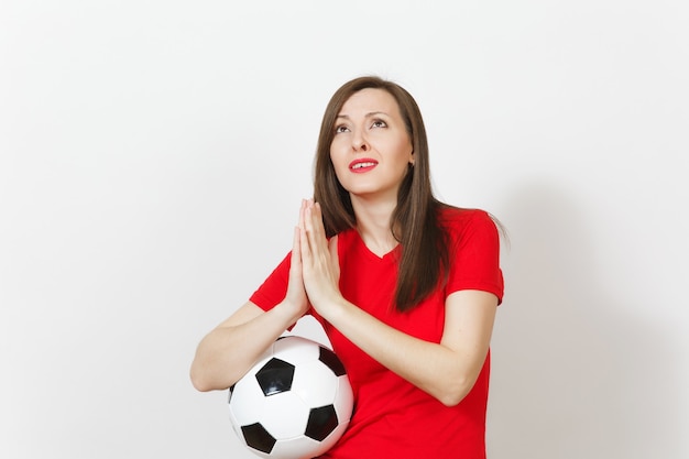 European young woman football fan or player in red uniform\
folded hands in prayer, hold soccer ball isolated on white\
background. sport play football lifestyle concept. wait special\
moment. make wish.