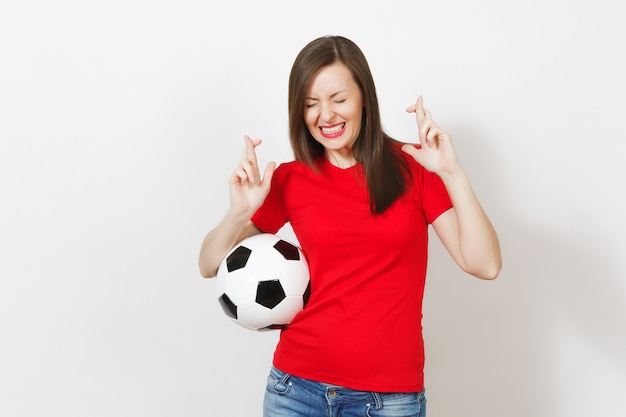 European young woman football fan or player in red uniform keep\
fingers crossed hold soccer ball isolated on white background.\
sport play football lifestyle concept. wait for special moment.\
make wish