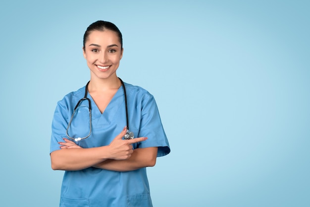 European young woman nurse in blue uniform pointing finger aside at copy space isolated on blue