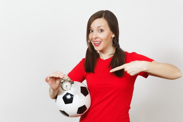 European young woman, two fun pony tails, football fan or\
player in red uniform hold old alarm clock, soccer ball isolated on\
white background. sport play football health, time lifestyle\
concept.