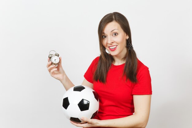 European young woman, two fun pony tails, football fan or\
player in red uniform hold old alarm clock, soccer ball isolated on\
white background. sport play football health, time lifestyle\
concept.