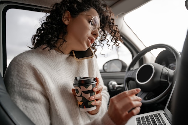 Europees meisje praten op mobiele telefoon in persoonlijke auto. ernstige jonge krullende zakenvrouw met gesloten ogen en het dragen van een bril. persoon met laptopcomputer en koffie papieren beker. multitasking