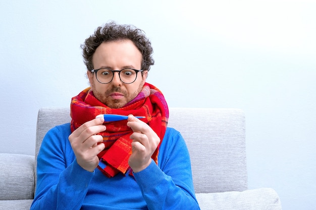 Foto europese geschokte mens die met rode sjaal temperatuur met digitale thermometerplaatsing thuis op bank meten. pandemische strijd, koorts genezen