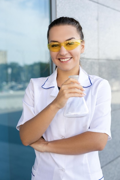 Europese glimlachende vrouwelijke wetenschapper in een witte jas en gele bril met een erlenmeyer