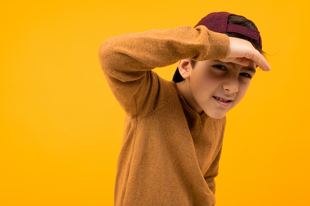 Europese knappe jongen tiener in een bruine trui en baseball cap is op zoek naar een doel terwijl hij zijn hand op zijn hoofd op geel houdt