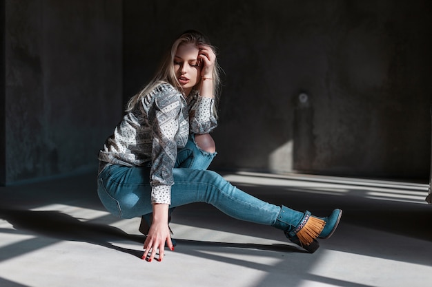 Europese modieuze jonge vrouw model in stijlvolle jeans in groene modieuze cowboylaarzen in een trendy shirt vormt zitten in een kamer met zonlicht