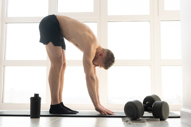 Europese sportman die zich uitstrekt op fitnessmat. Zijaanzicht van jonge, bebaarde man met naakte torso. Concept van sportactiviteit thuis. Interieur van modern ruim appartement. Zonnige dag