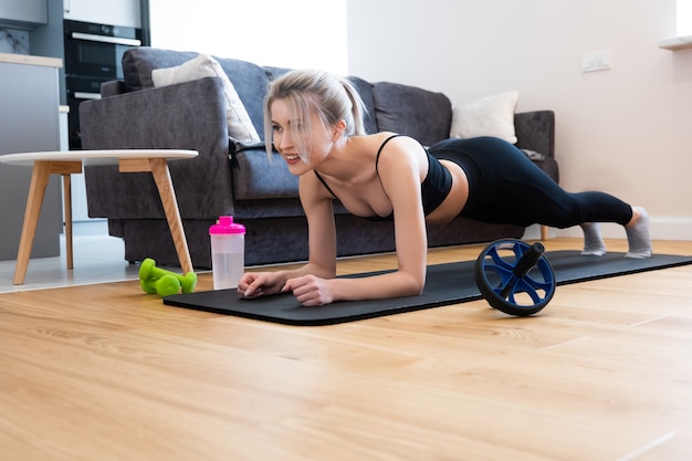 Europese sportvrouw doet plank voor buikspieren op fitnessmat. Jonge mooie lachende blonde vrouw draagt sportkleding. Concept van sportactiviteit thuis. Interieur van studio appartement