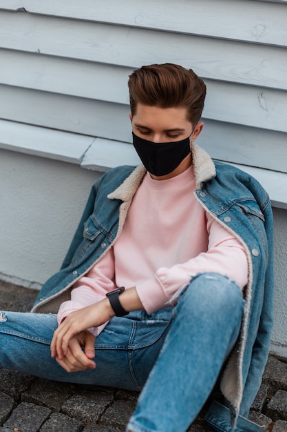 Europese stijlvolle jongeman in denim modieuze blauwe vrijetijdskleding in beschermend zwart masker rust in de buurt van het voortbouwen op straat in de stad. Urban fashion model guy is beschermd tegen coronavirus. Stijl 2020.