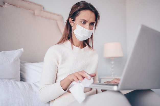 Europese vrouw in gezichtsmasker in slaapkamer tijdens coronavirus isolatie huis quarantaine schoonmaak laptop