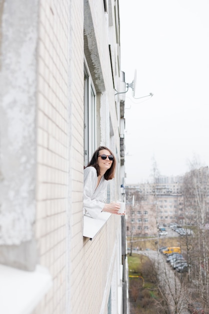 Europese vrouw in zonnebril en met een mok koffie of thee die uit het raam kijkt en glimlacht