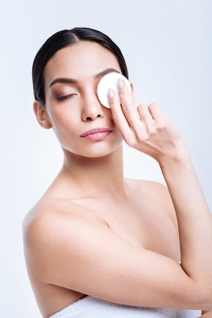 Evening routine. Charming dark-haired woman removing her eye makeup with a cotton pad before going to shower while being wrapped up in a towel