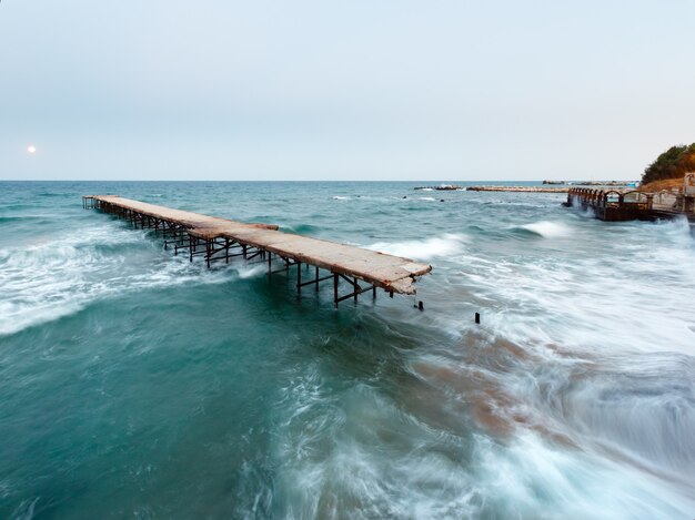 Evening sea surf view, ruined pier and Moon in sky (Black Sea, Bulgaria). Long-time exposition