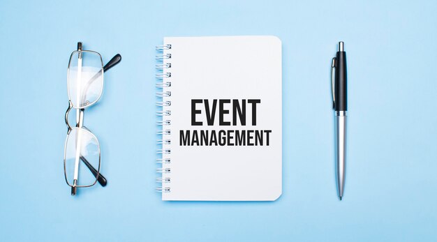 Event Management words in white notepad, pen and glasses on blue background. Concept