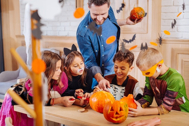 Event manager. Bearded handsome event manager helping children wearing Halloween costumes decorating pumpkins