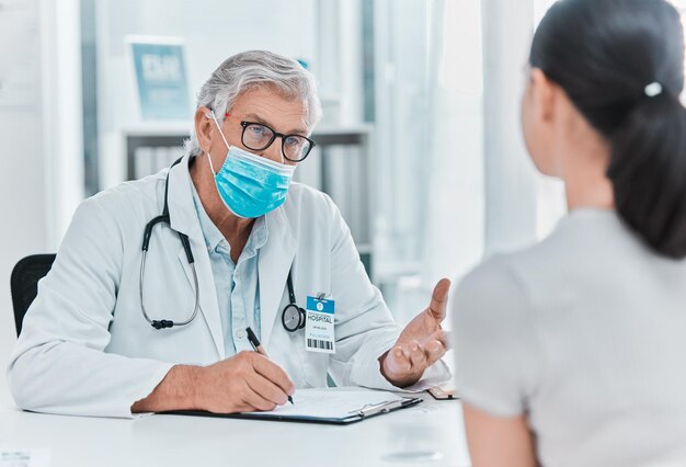 Eventuele hoofdpijn Shot van een volwassen arts die een gezichtsmasker draagt en bij zijn patiënt zit tijdens een consult in zijn kliniek