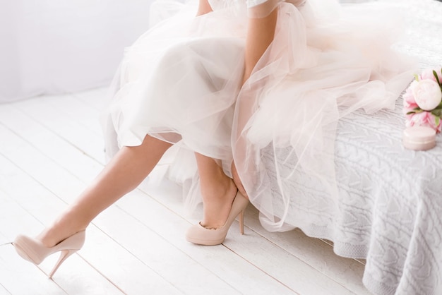 Every detail is important. Charming pleasant young bride sitting on the bed in white colored room while wearing her shoes and expressing tenderness