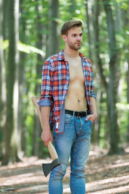 Every man needs some time for himself Handsome guy hold axe in woods Unshaven guy with sexy torso in open shirt Lumberjack style Summer vacation Natural landscape Traveling and wanderlust