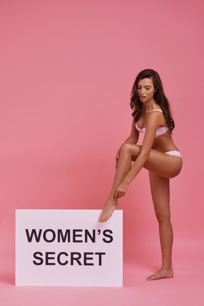 Every woman is a mystery.  Full length of attractive young woman touching her leg while standing against pink background