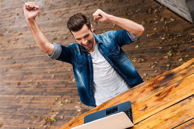 Everyday winner. Top view of excited young man keeping arms raised and expressing positivity while sitting at the wooden table outdoors