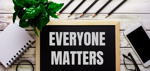 EVERYONE MATTERS is written in white on a black board next to a phone, notepad, glasses, pencils and a green plant