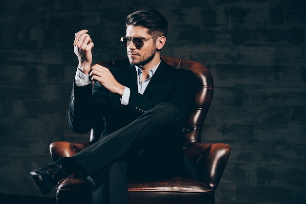 Photo everything must be perfect.young handsome man in suit and sunglasses adjusting sleeve on his shirt while sitting in leather chair against dark grey background