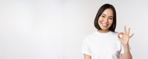 Everything okay Smiling young asian woman assuring showing ok sign with satisfied face standing over white background Copy space