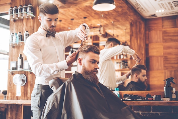 Tutto dovrebbe essere perfetto. vista laterale del giovane uomo barbuto che si fa tagliare i capelli dal parrucchiere mentre è seduto in poltrona al barbiere