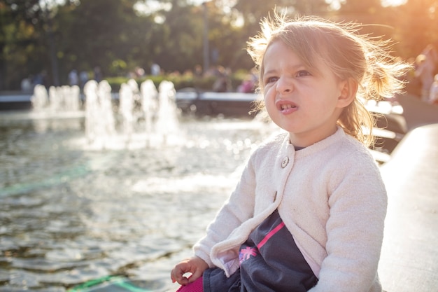 Evil expression face. Portrait of little angry toddler girl she grimaces and shows a grin in the sunny park outdoors